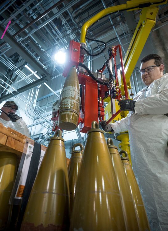 Person wearing safety gear operating machinery handling artillery shells.