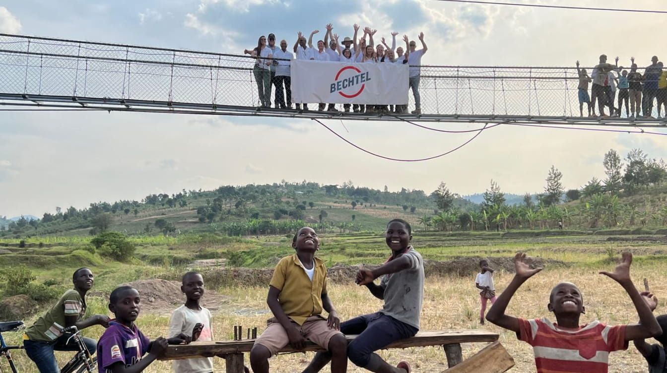 Group of bechtel.org volunteers waving from suspension trail bridge holding a Bechtel banner, with young community members.