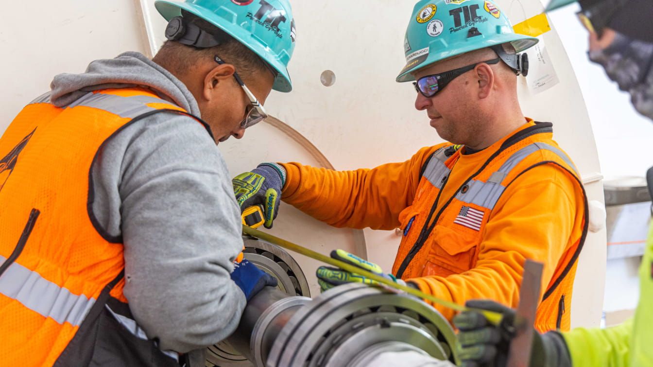 Two people wearing safety gear measuring a pipe.