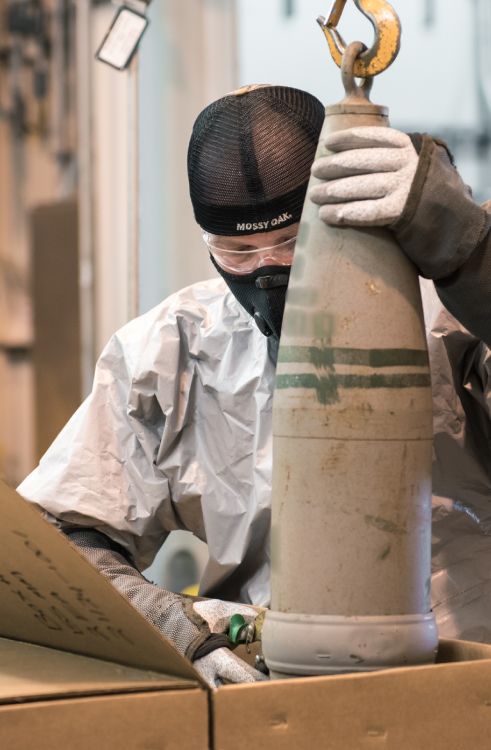 Person in safety gear lifting a large artillery shell out of a box.