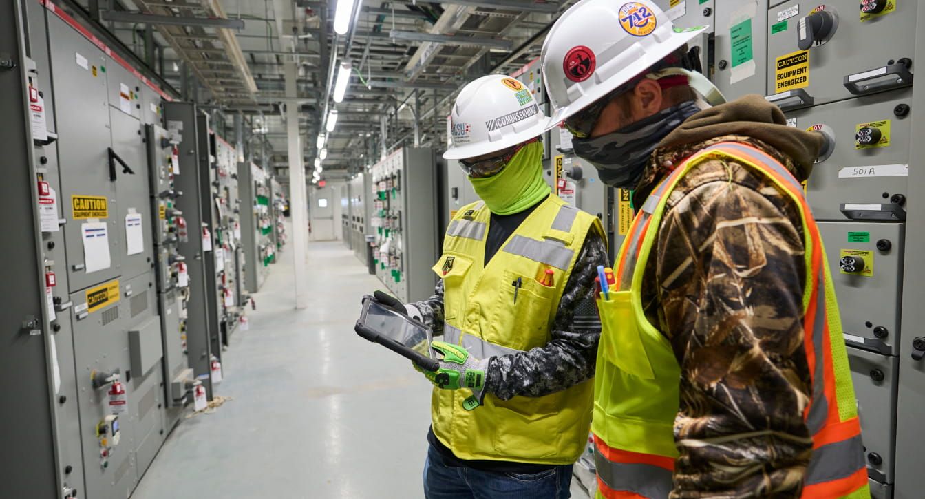 Bechtel team members wearing safety gear standing in an electrical room, looking at a tablet.