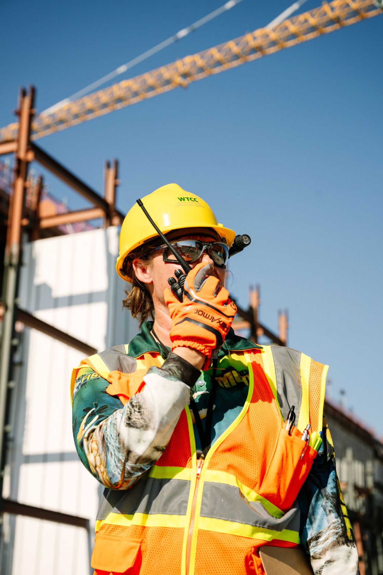 Woman in a hard hat and safety vest on project site talking into a handheld radio.