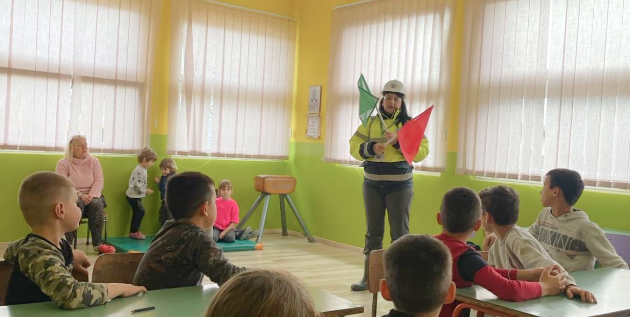 Person wearing safety gear, holding two safety flags during a presentation to young students in classroom.