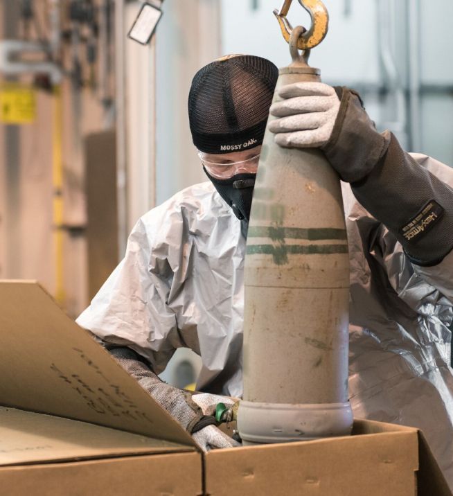 Person in safety gear lifting a large artillery shell out of a box.