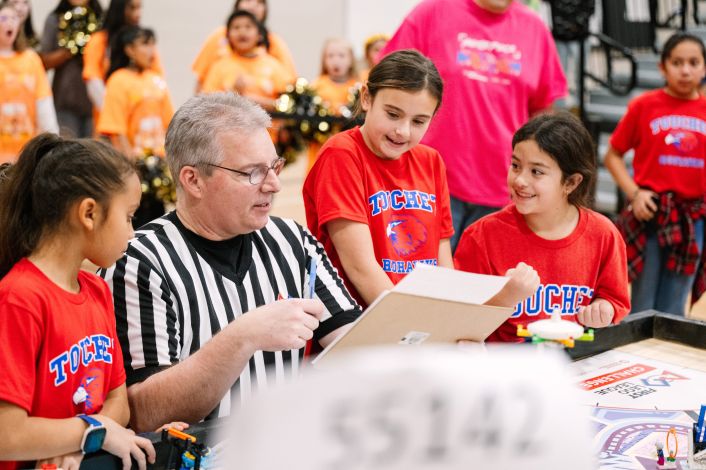 A man in a referee uniform talking to some children in red school sports shirts.