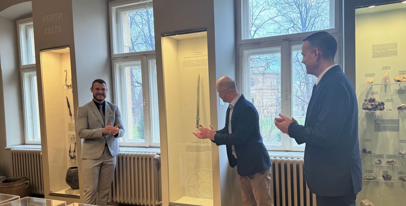 Three men clapping in front of an antique sword at a sword museum.